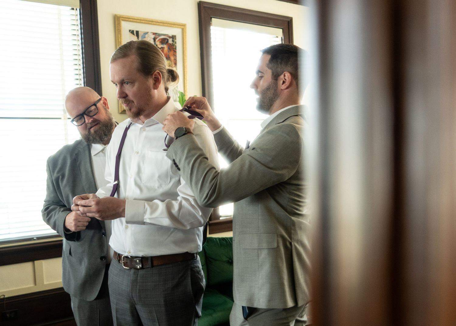 A group of men in business attire interact during a formal fitting or preparation session at an indoor venue.