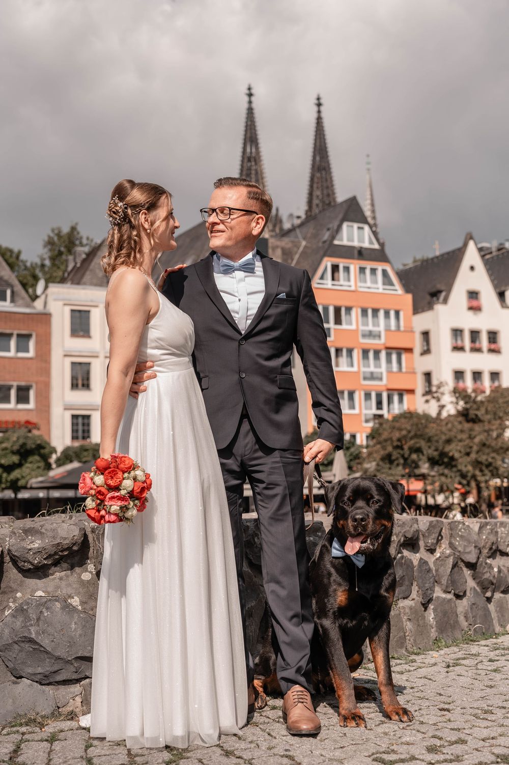 A wedding couple poses with their black dog against a backdrop of Gothic church spires in a European city.