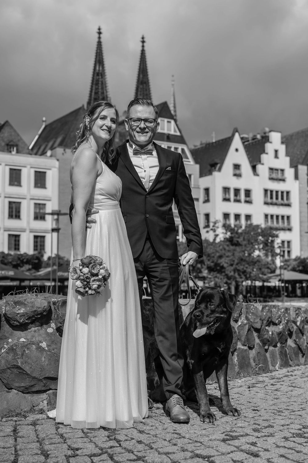 A black and white wedding portrait featuring a couple and their dog in front of historic European architecture.