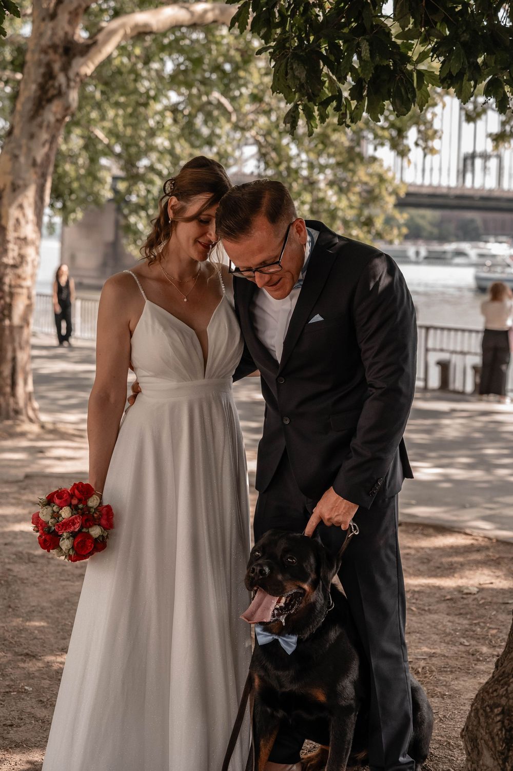 A sunny outdoor wedding portrait by the river with a black dog joining the celebration.