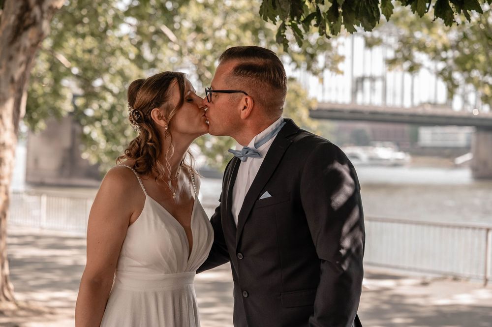 A bride and groom share a romantic kiss in a sun-dappled outdoor setting with trees in the background.
