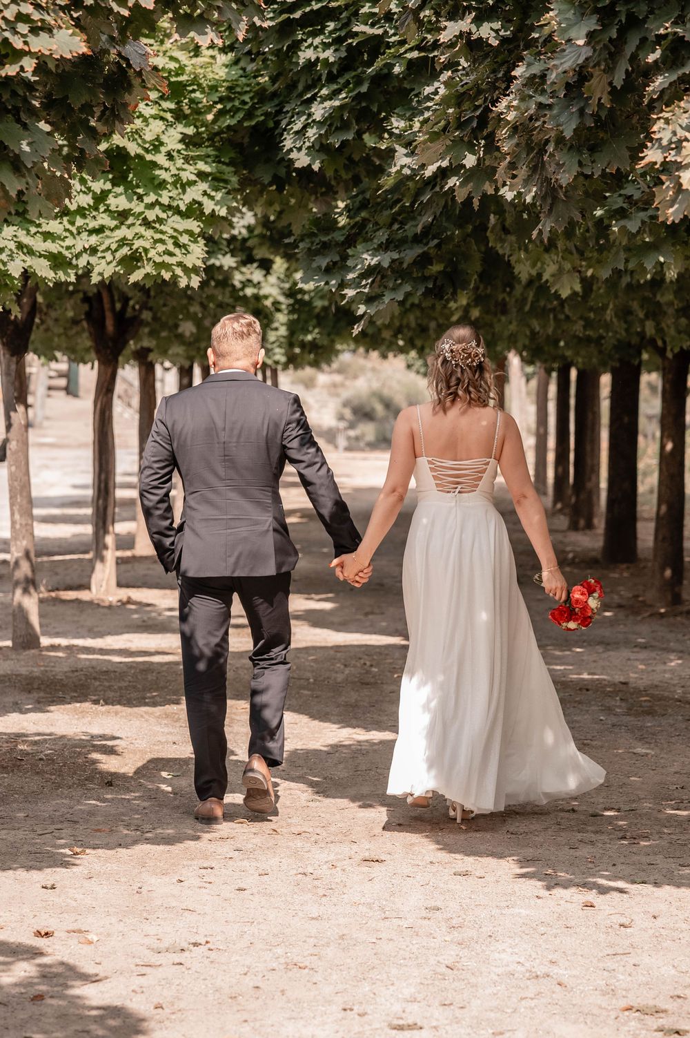 A couple in wedding attire walks away from the camera holding hands beneath a tree-lined path in a romantic woodland setting.