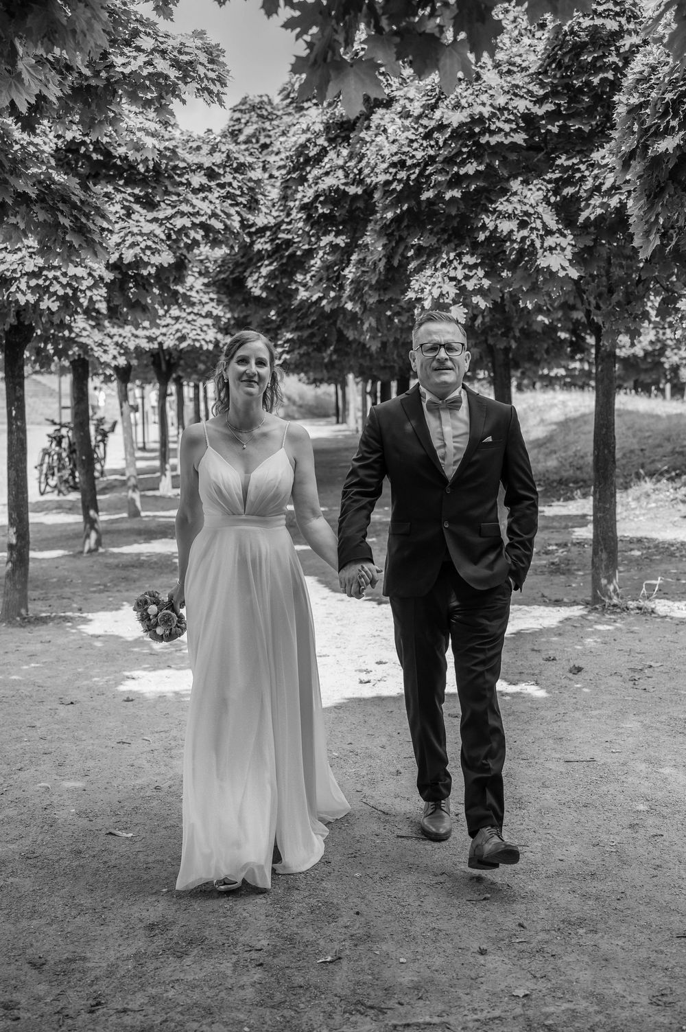 A black and white wedding photo of a couple walking together through a tree-lined path in formal attire.