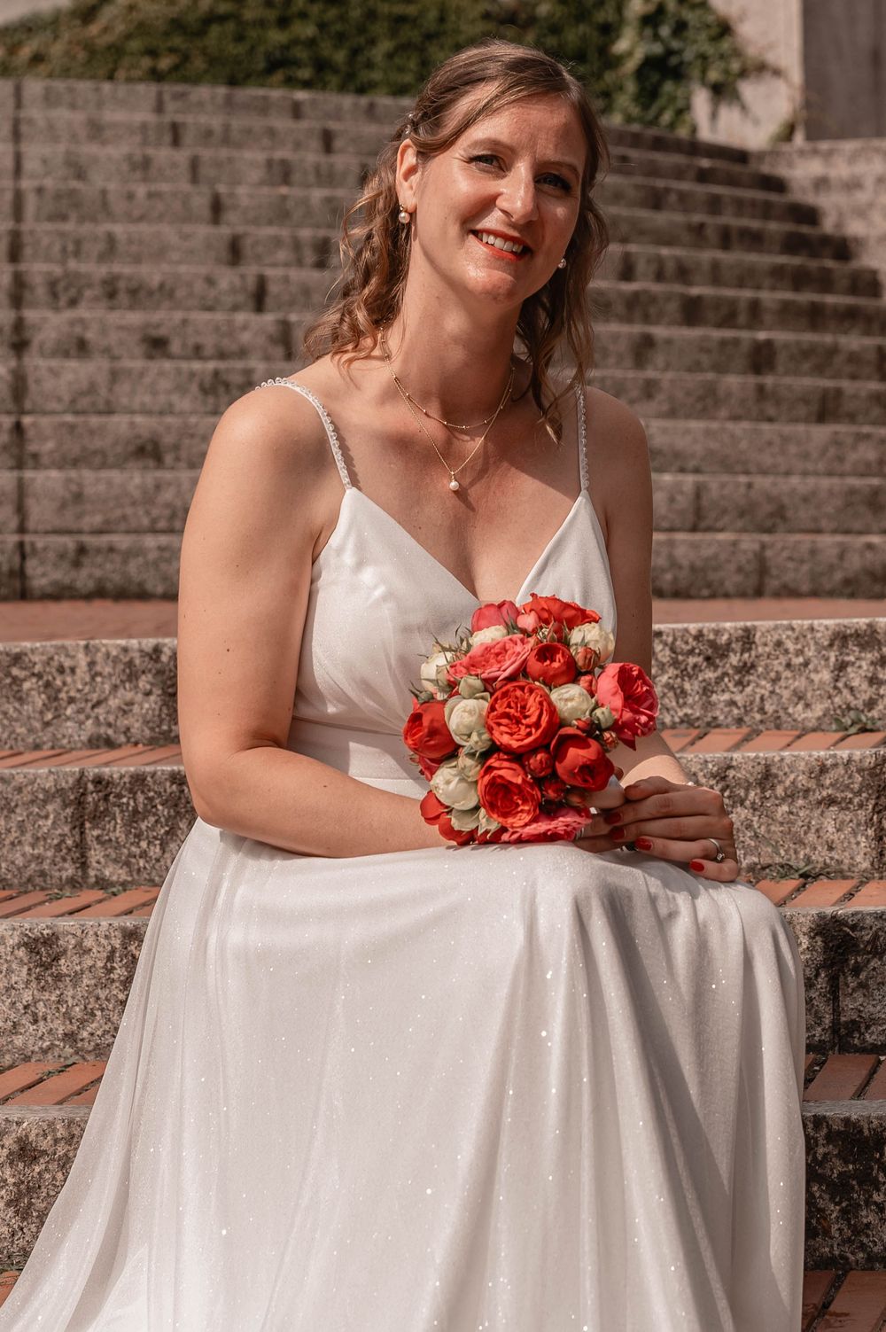 A bride in a flowing white dress poses on stone steps while holding a red rose bouquet.