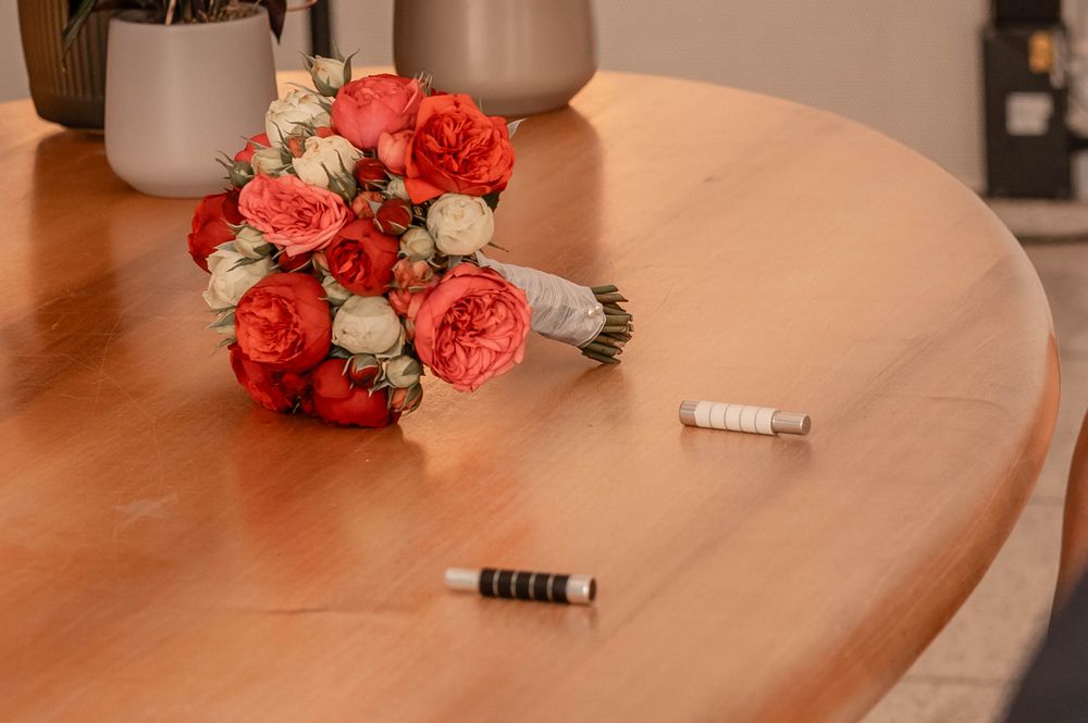 A coral pink and white rose bouquet rests on a wooden table beside two small cylindrical objects.