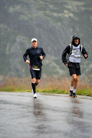 Two athletes running on a wet mountain road wearing race bibs and athletic gear during competition.