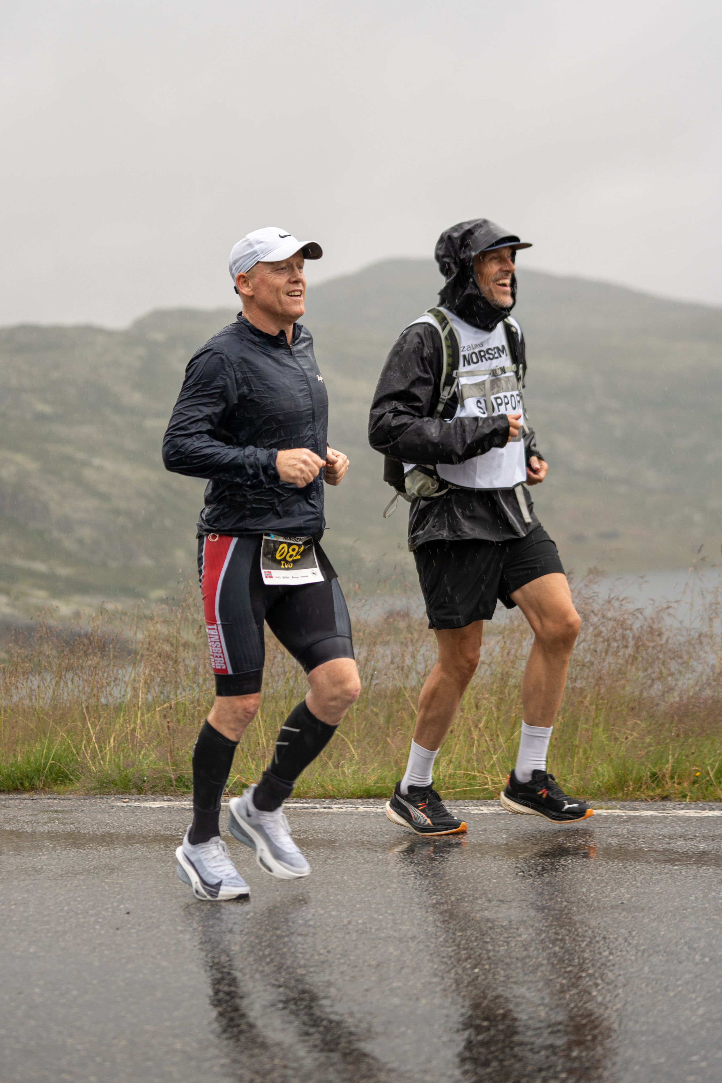 Two athletes run together on a wet road during an endurance race in misty mountain conditions.