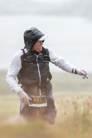 A person in a dark vest and rain hat leans into strong wind while standing in a misty grass field.