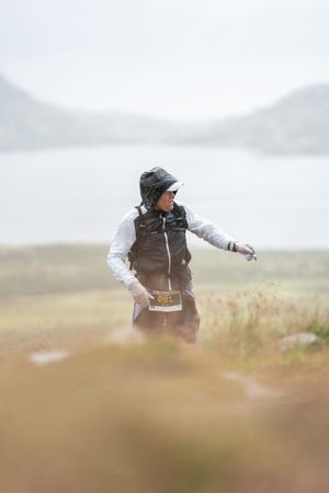 Athlete in windbreaker and hat photographed through tall grass against an overcast mountain landscape.