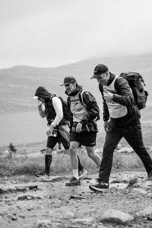 Three hikers with backpacks traverse rocky mountain trail in black and white.