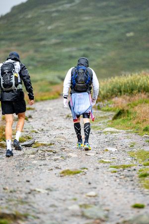Two trail runners with backpacks traverse rocky mountain path during endurance race.
