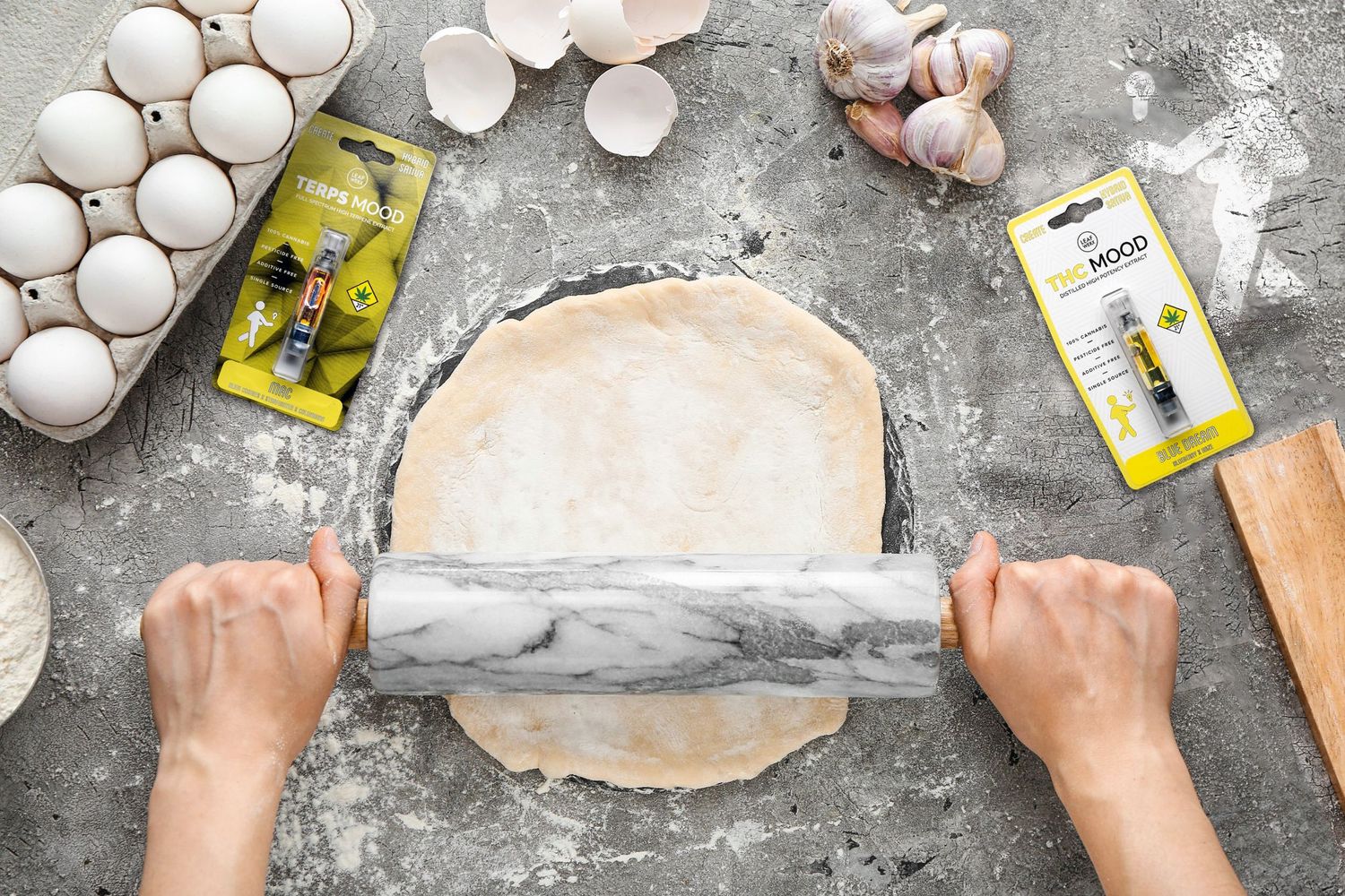 Hands rolling out pizza dough on a marble board with flour, eggs, and yeast packets on a concrete surface.