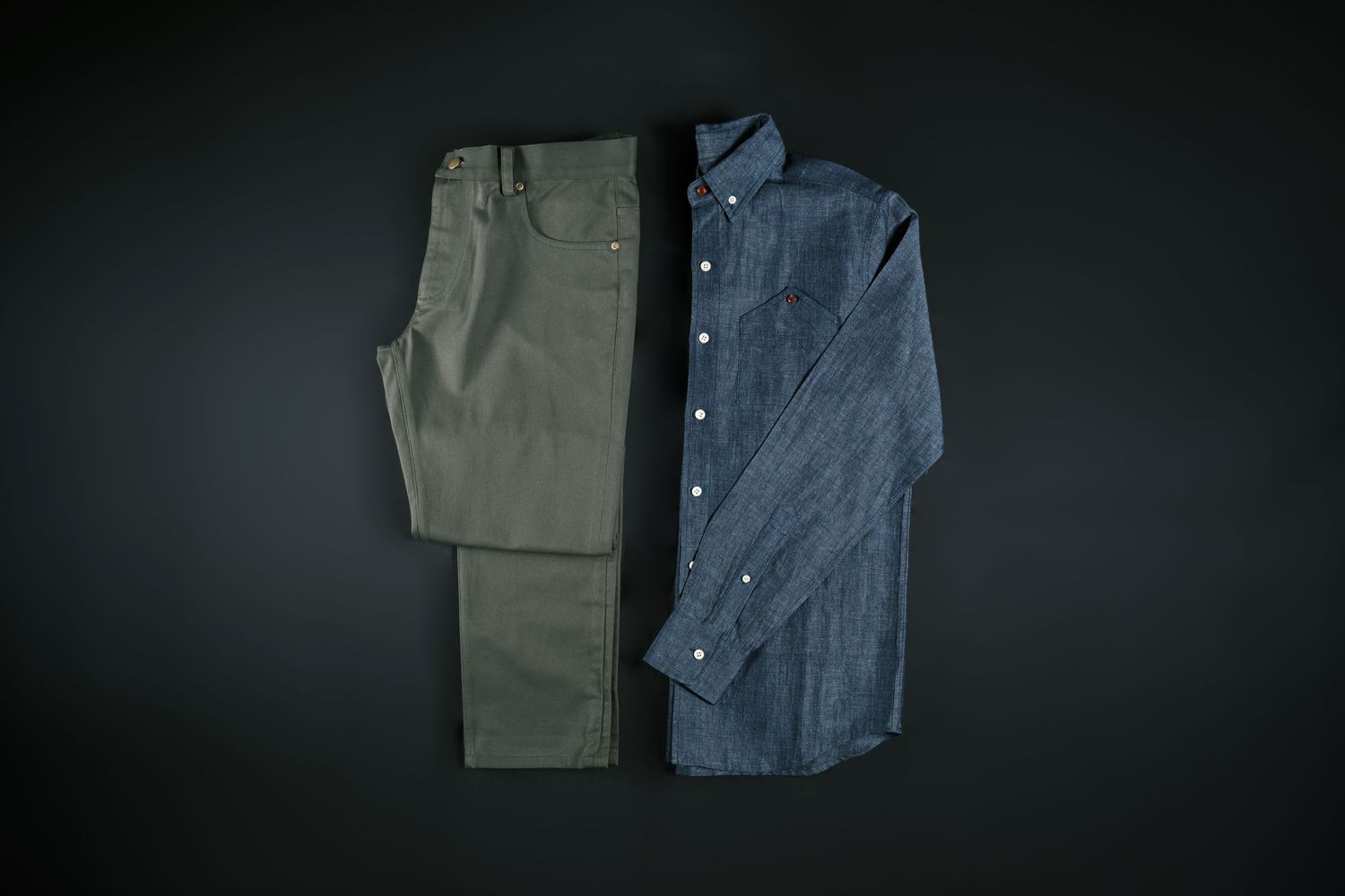 A flat lay of a chambray button-down shirt paired with olive green chino pants on a dark background.