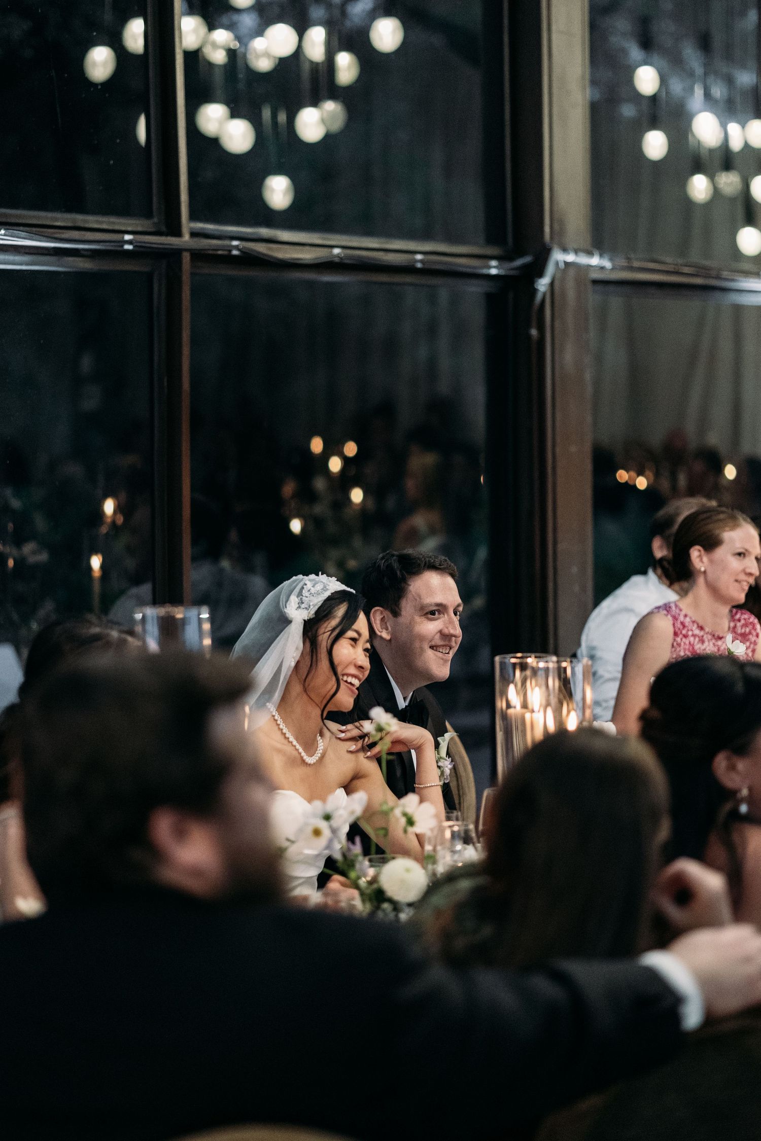Wedding guests seated at a candlelit reception dinner smile and laugh while enjoying the evening festivities.