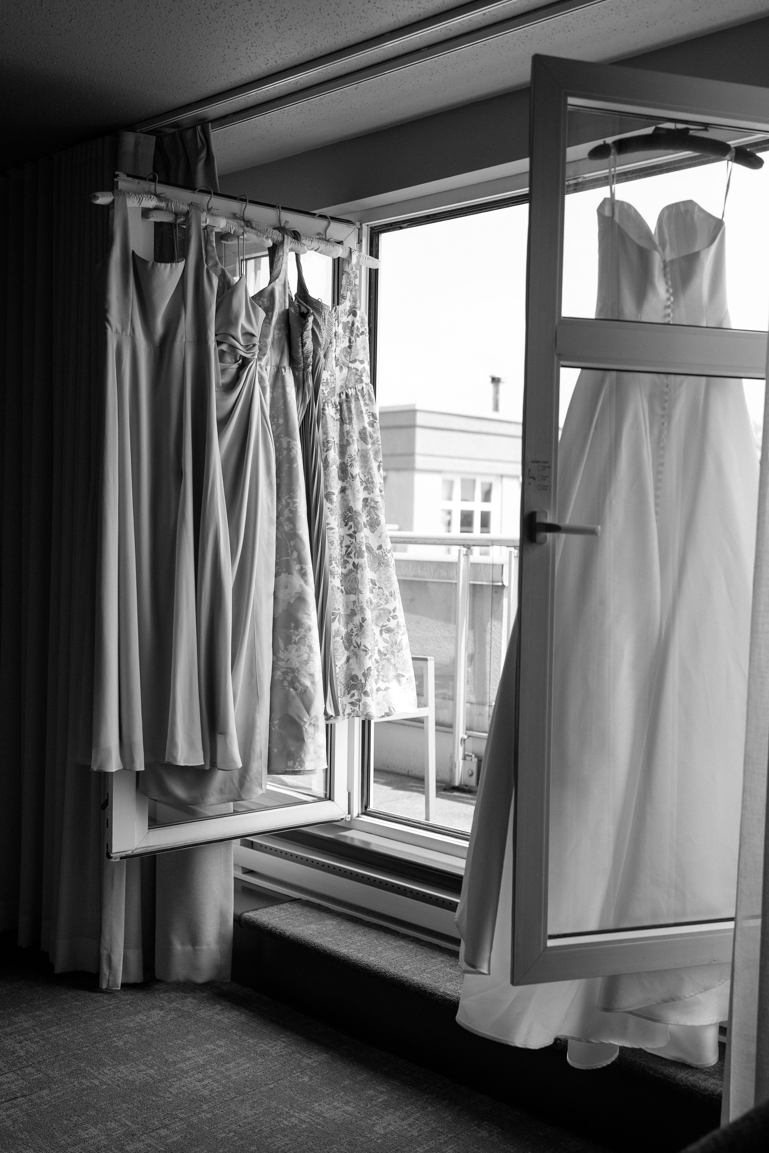 Black and white photo of elegant wedding dresses and bridesmaid gowns hanging by a sunlit window.