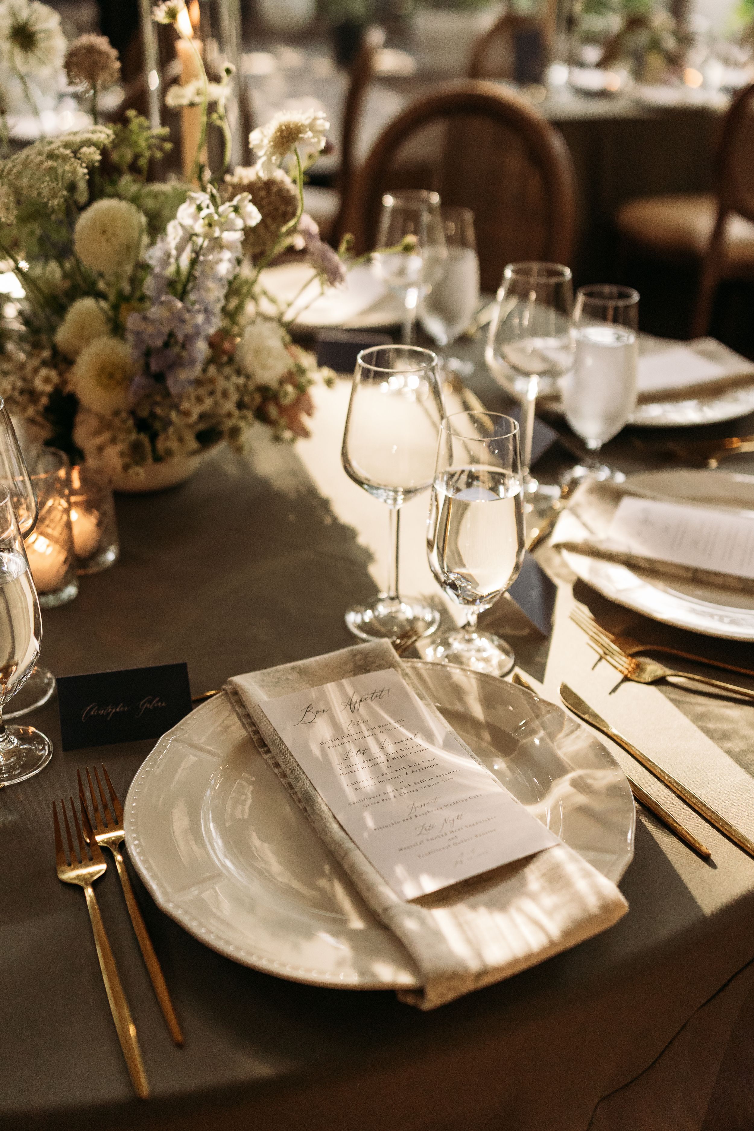 Elegant wedding table setting with gold cutlery, white plates, wine glasses and floral centerpiece on dark tablecloth.