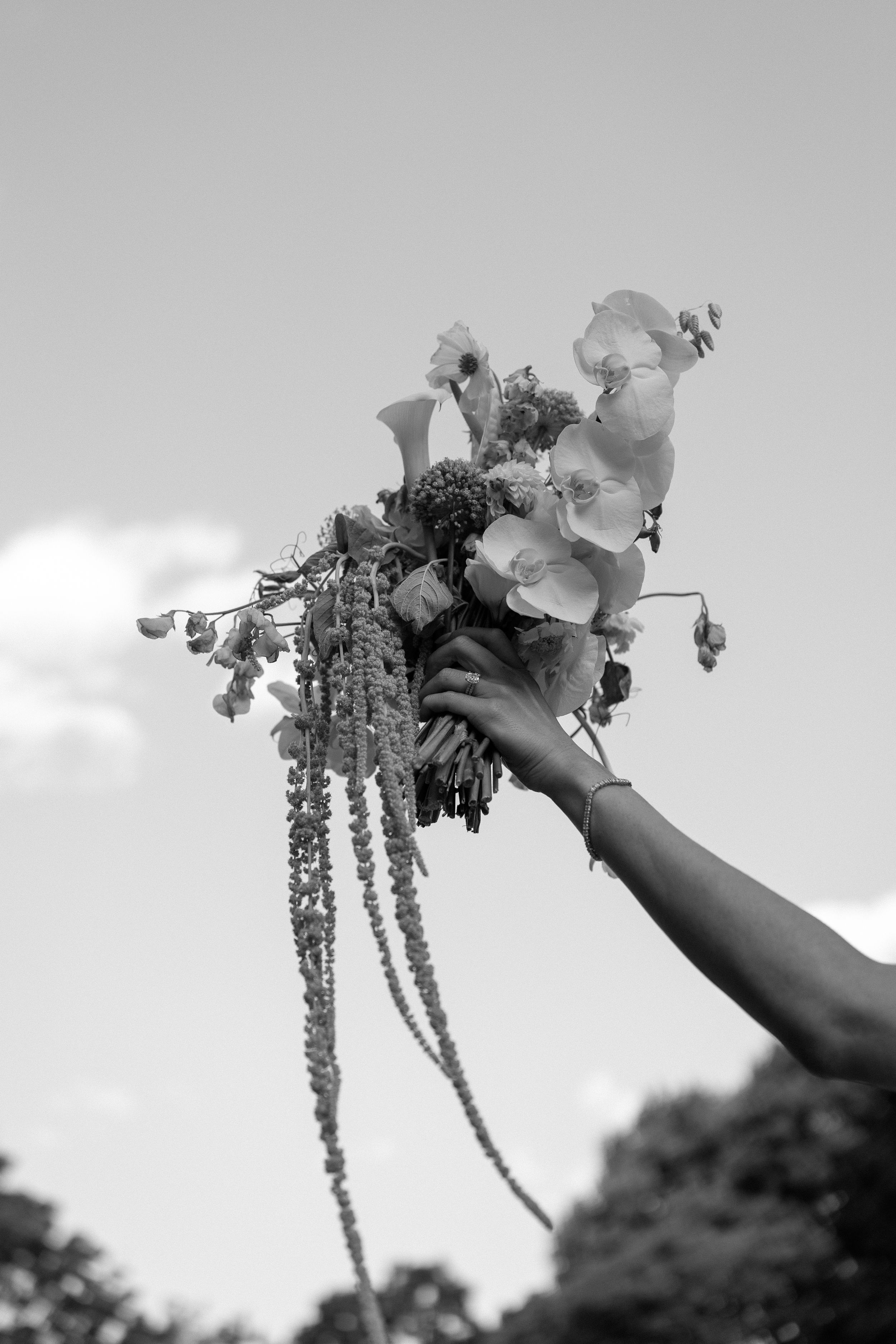 Dramatic black and white photo of a cascading bridal bouquet with orchids held against a bright sky.
