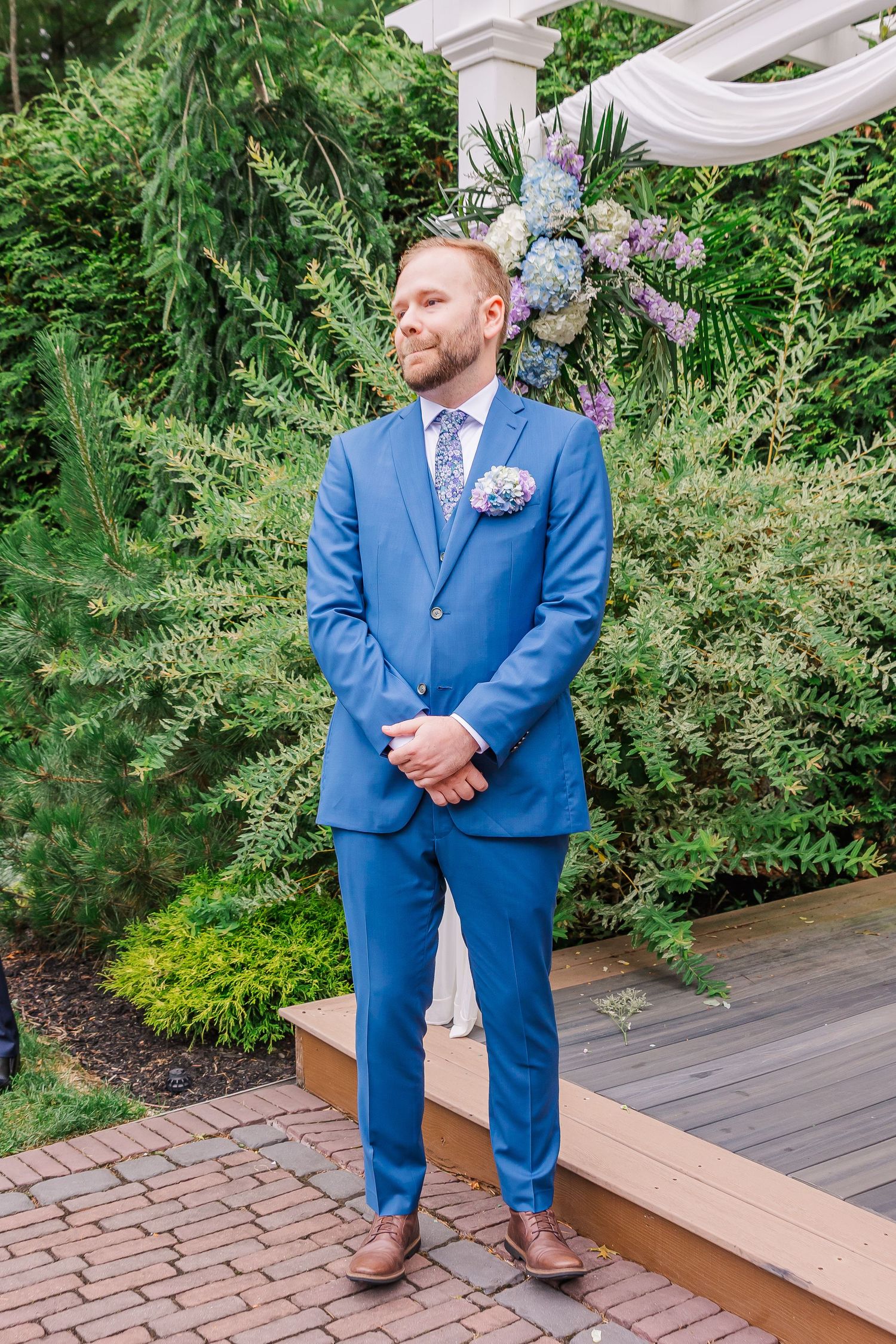 Wedding guest in a purple lace-detailed formal dress stands by white chairs decorated with floral arrangements on brick patio.