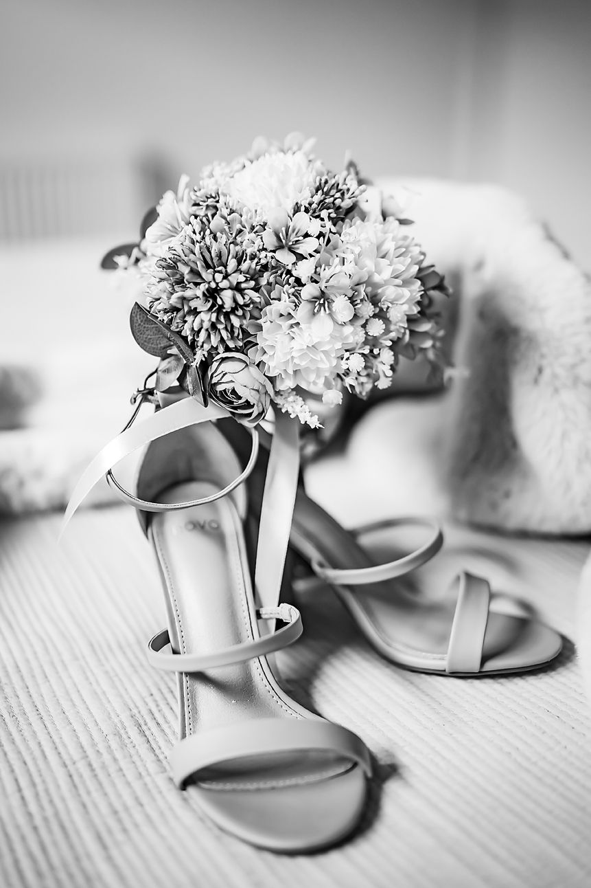 Black and white wedding photo of elegant bridal heels with floral bouquet containing white flowers and greenery.