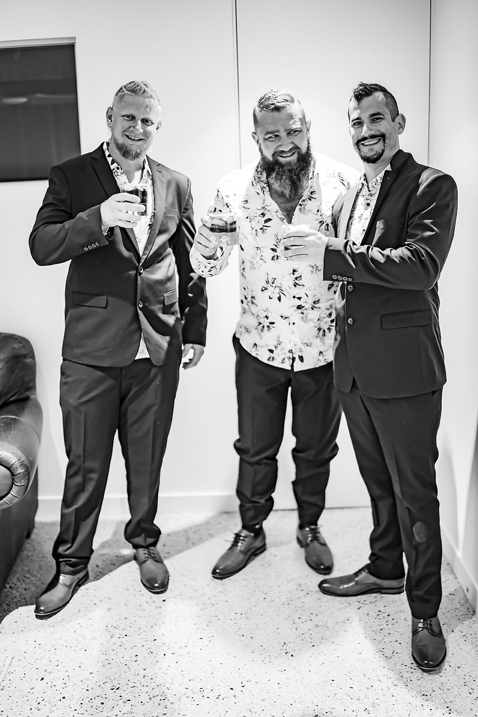 Three men in formal attire stand together smiling and holding drinks at an indoor event in black and white.