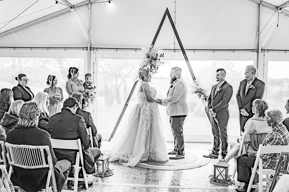 A wedding ceremony takes place under a triangular arch inside a white tent with guests seated in folding chairs.