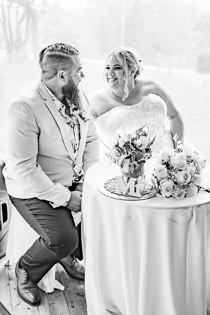 Black and white candid wedding photo showing a couple sharing a joyful moment beside a table with floral centerpiece.