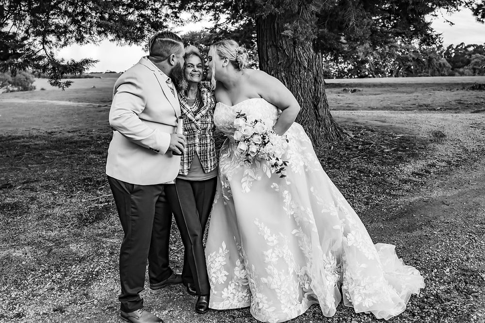 Black and white outdoor wedding photograph showing a joyful moment between people under a tree with the bride in a flowing dress.