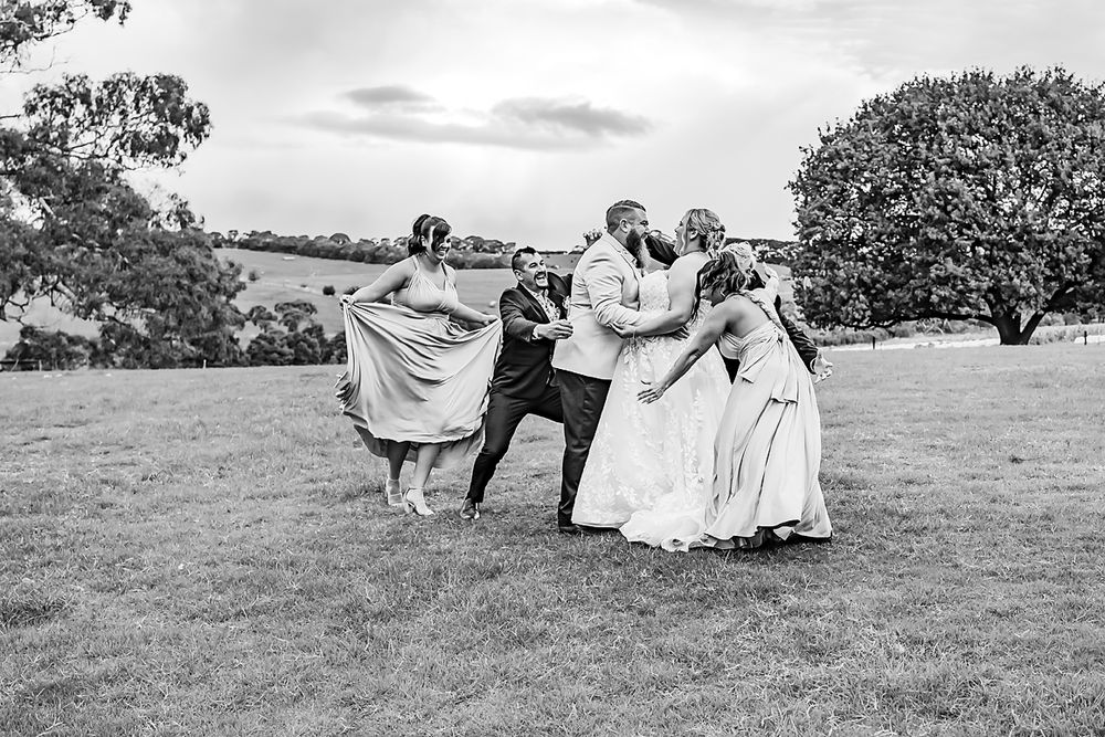 A joyful wedding party holding dresses and celebrating together on a grassy hill in black and white photography.