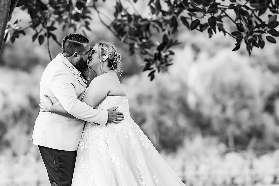 A romantic black and white wedding photo shows a couple embracing under tree branches in a dreamy outdoor setting.