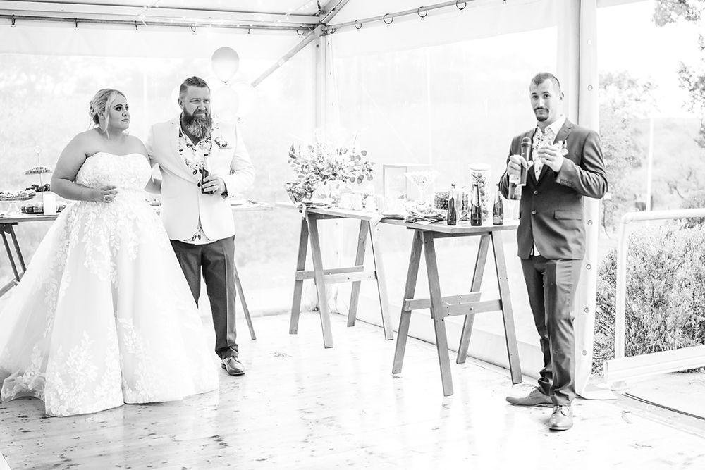 Black and white photo of guests enjoying speeches during an outdoor wedding reception under a tent.