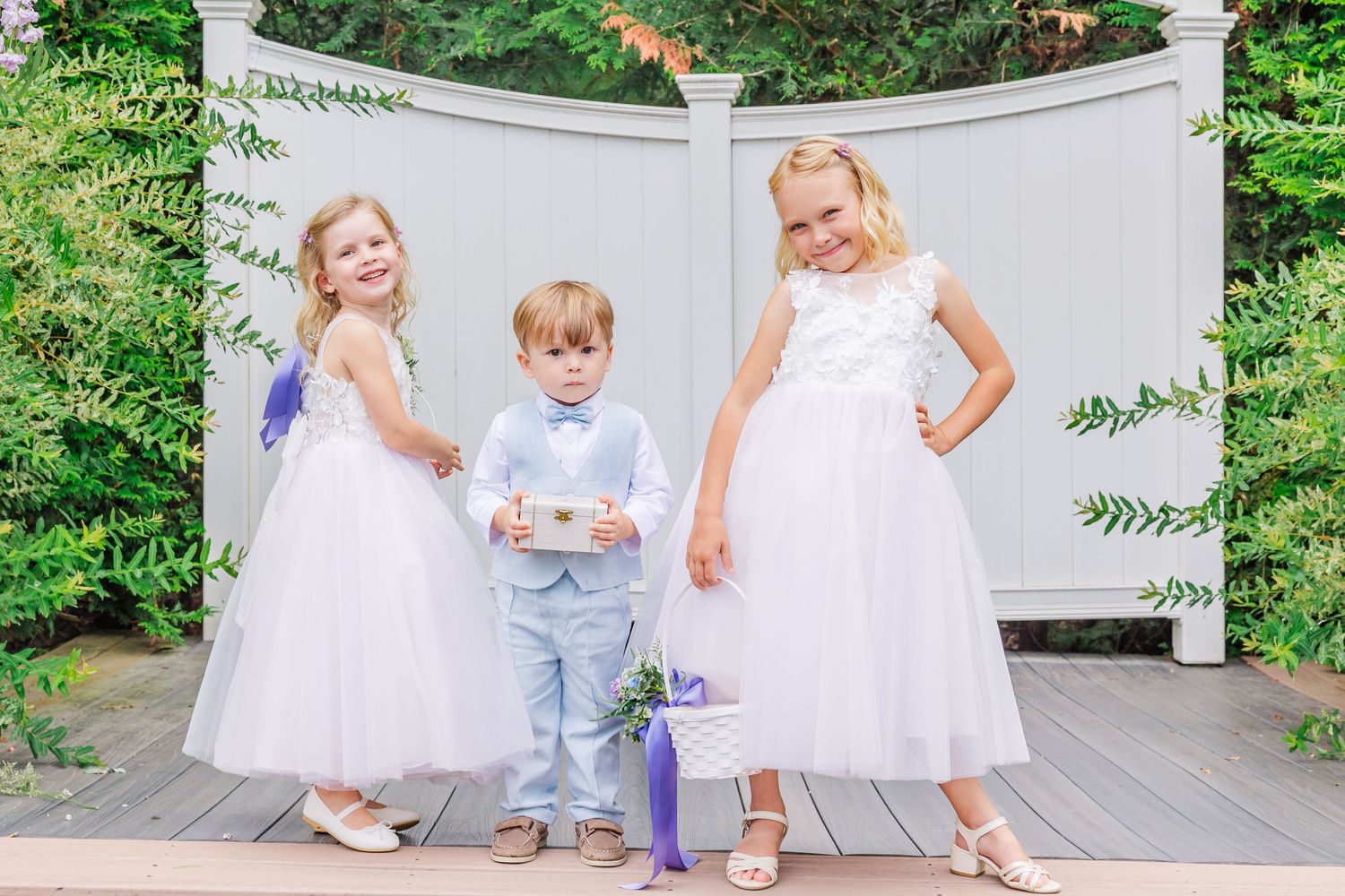 Three toddlers in white dresses dance and play together on a wooden deck in a garden setting.