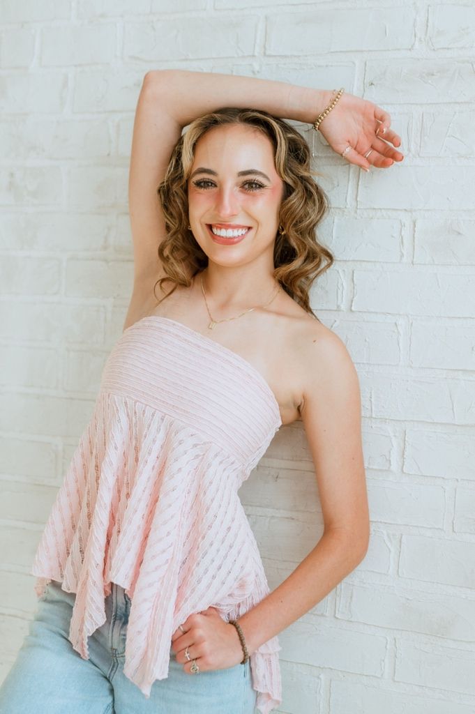 A person in a pink strapless top poses against a white brick wall with one arm raised and curled hair.