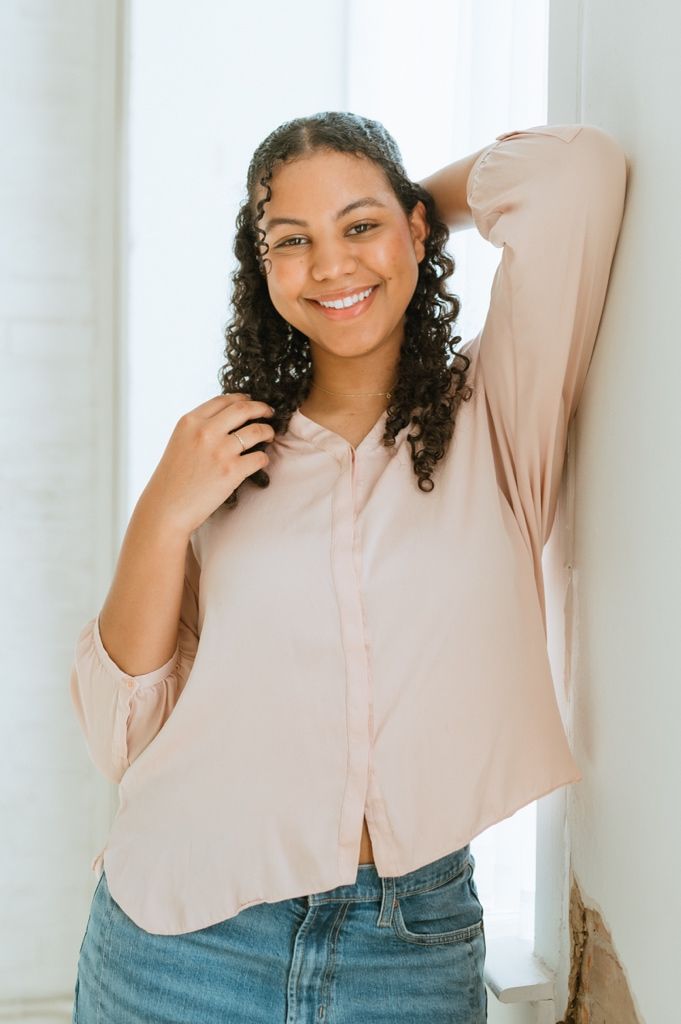 Young woman wearing a pale pink button-up blouse and jeans poses casually by a white curtain with natural curly hair.