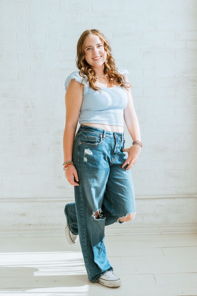 A person in a light blue crop top and distressed baggy jeans poses casually against a white wall.