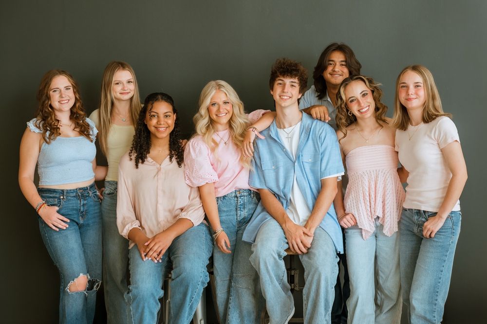Group of young people wearing casual denim and pastel tops pose together against a teal colored backdrop.