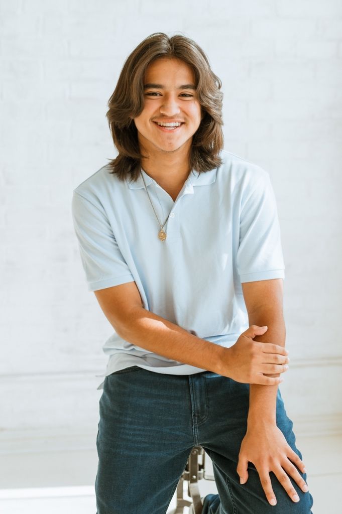 Young person with shoulder-length hair wearing a white polo shirt and jeans smiles against a light background.