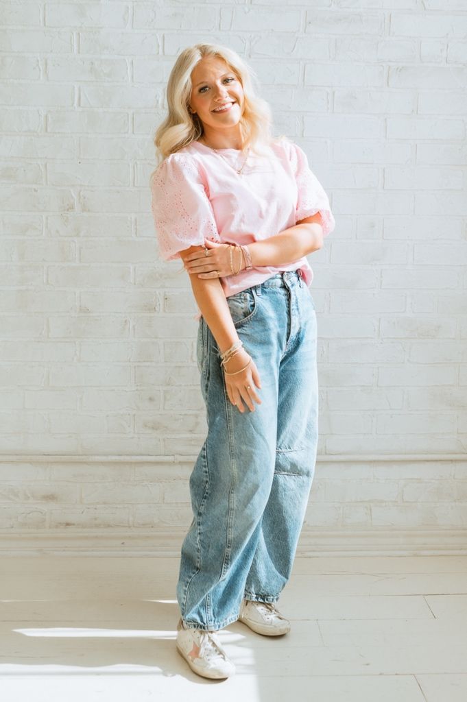 A person in a pink sweater and baggy light wash jeans poses casually against a white brick wall.