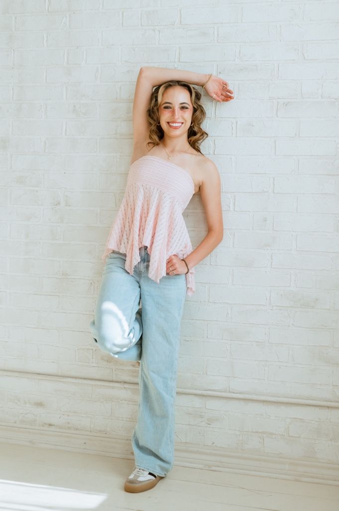 A person in a pink sleeveless top and light blue jeans poses casually against a white brick wall.