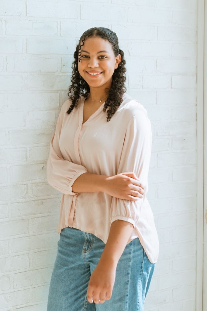 A person in a blush-colored blouse and denim poses warmly against a white brick wall background.