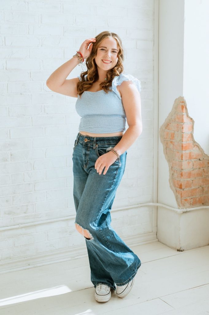 Person in light blue top and baggy jeans poses playfully against white wall in bright minimalist setting.