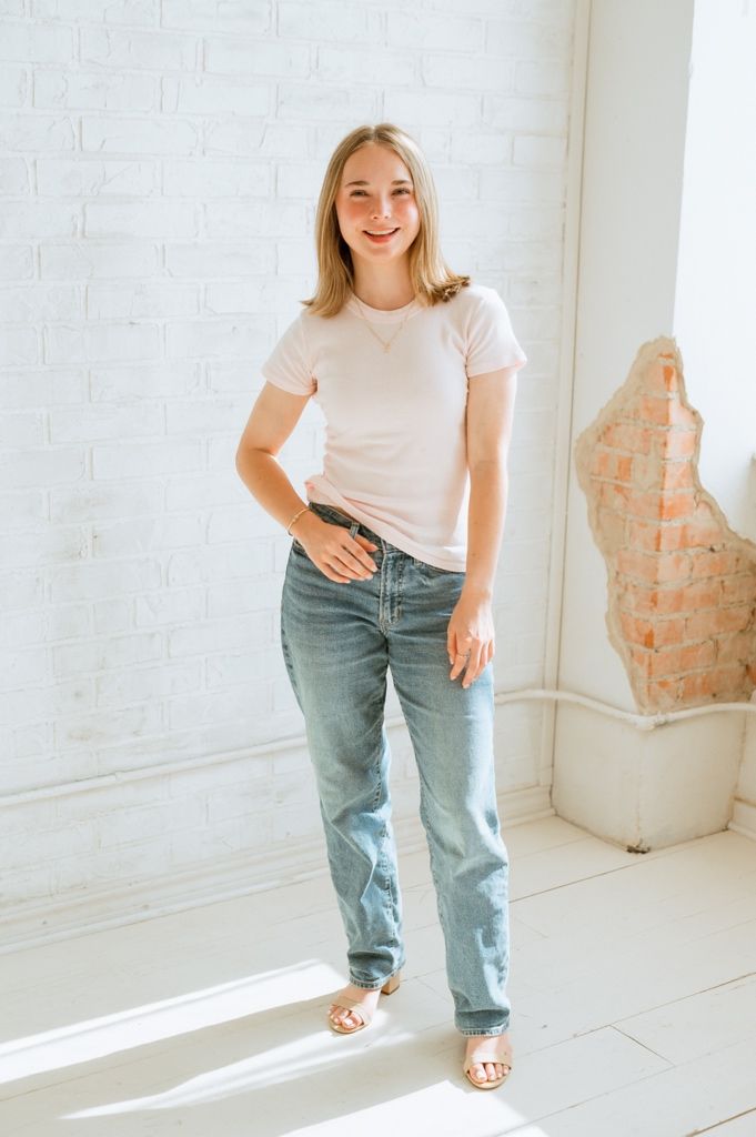 A person in a white t-shirt and baggy jeans poses casually in a bright minimalist room with bare brick walls.