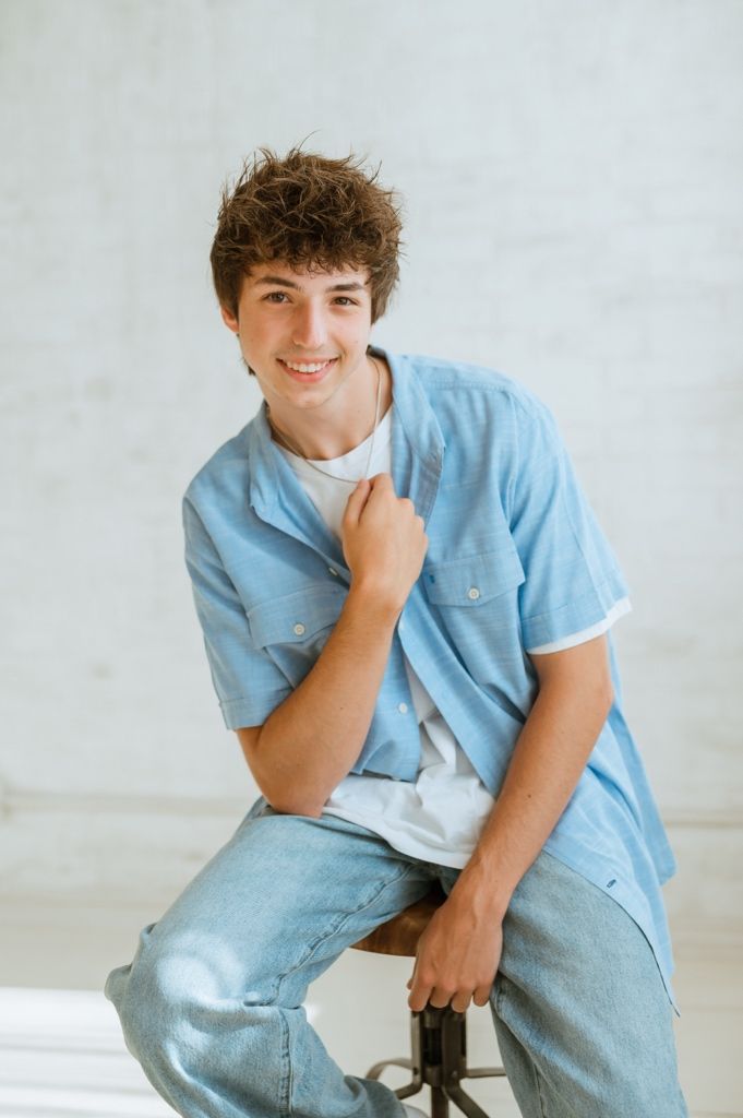 Young person in light blue button-down shirt and jeans sits casually on a stool against white backdrop.