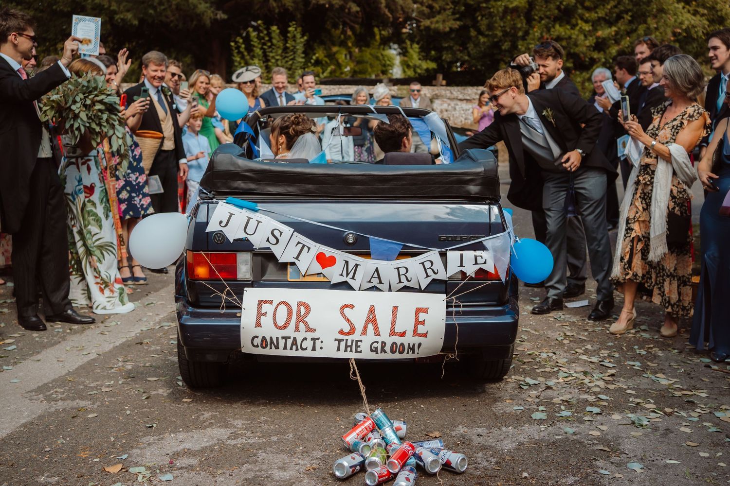 A wedding party surrounds a decorated car with 'Just Married' and 'For Sale' signs during an outdoor celebration.
