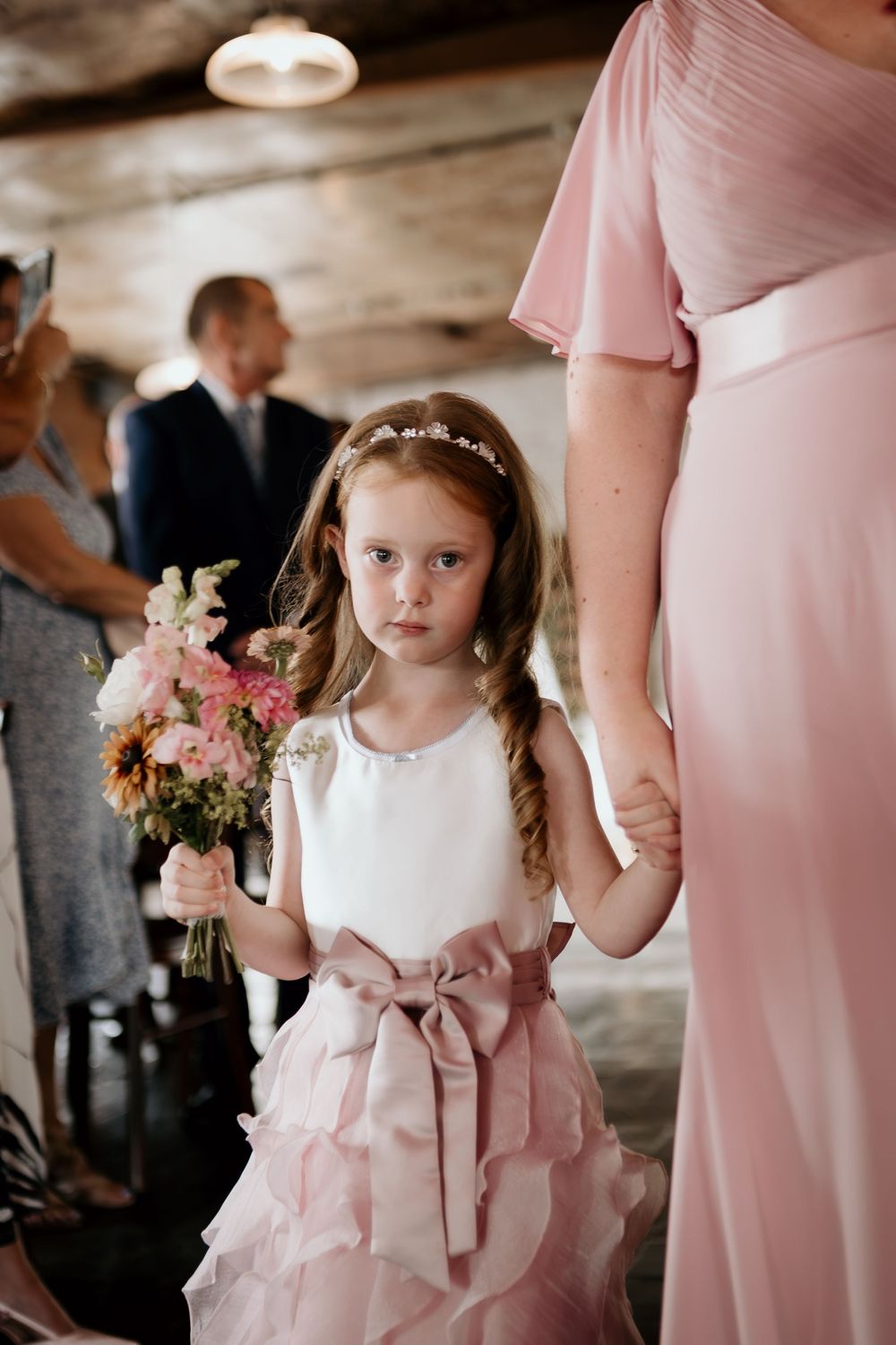Young flower girl in white and pink dress holding bouquet at wedding ceremony.