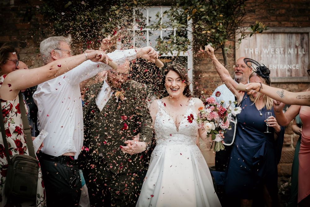 Wedding party showering newlyweds with confetti against brick wall backdrop.