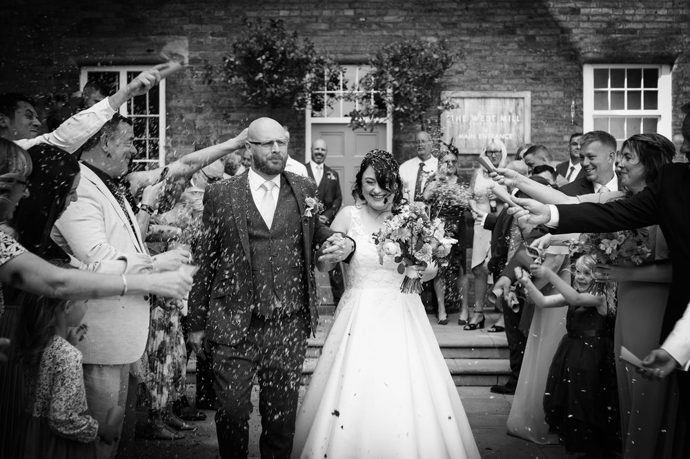 Joyful wedding celebration with guests throwing confetti at a couple exiting a building in black and white photography.