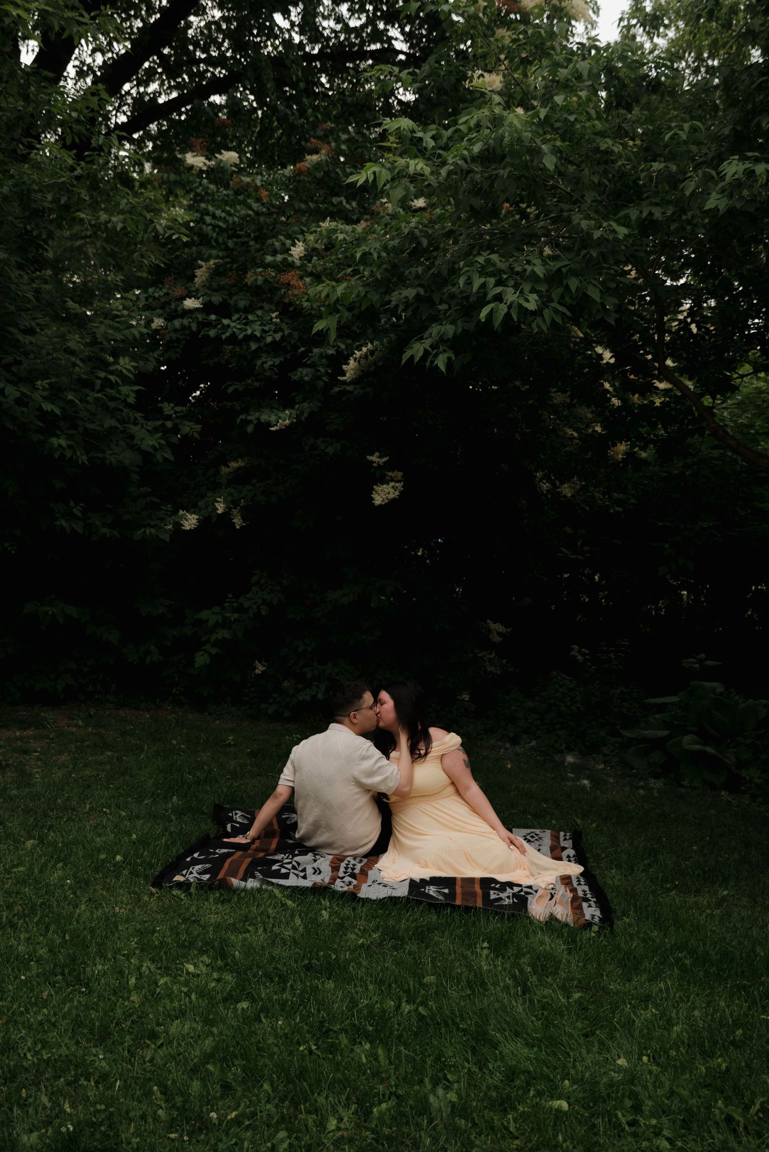 A couple sits on a blanket for a romantic picnic in a lush green backyard surrounded by trees at dusk.