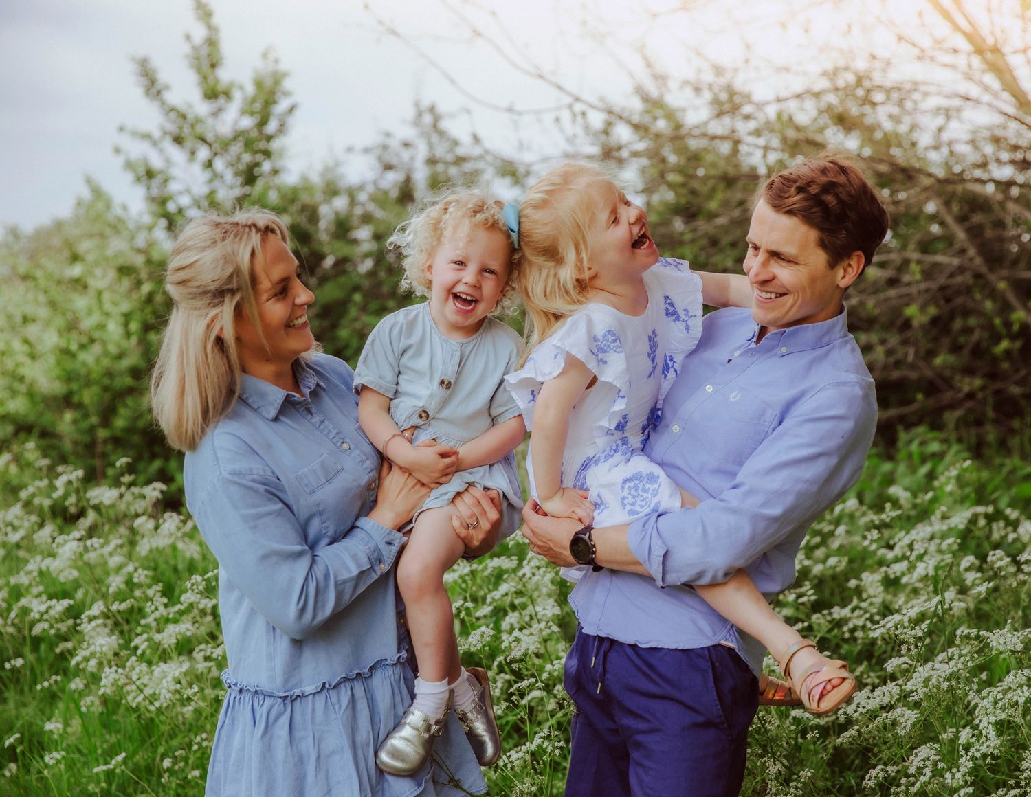 A family wearing coordinated blue outfits enjoys a playful moment together in a blooming wildflower meadow. During a fun family photoshoot