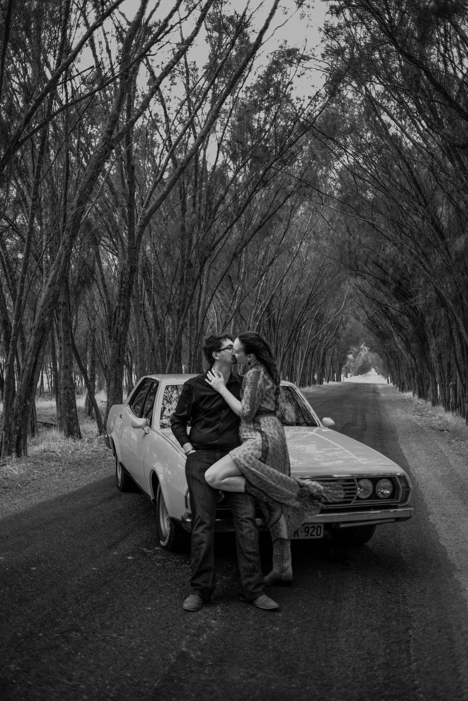 A tender romantic moment captured in black and white as a couple embraces on the hood of a classic car.