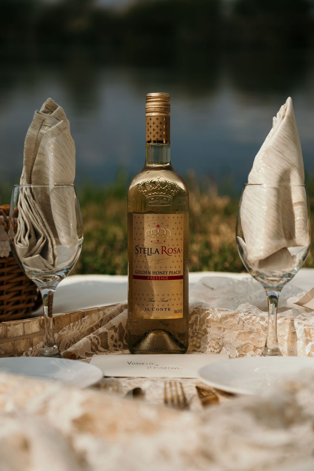 A bottle of Stella Rosa wine stands between two champagne glasses on an outdoor picnic table near a lake.